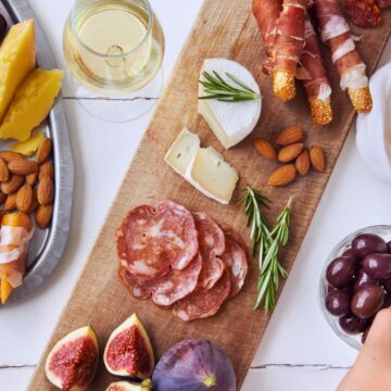 A charcuterie board with assorted cheeses, salami, prosciutto-wrapped breadsticks, nuts, figs, and fresh rosemary. Accompanied by grapes, a loaf of bread, and a side dish of mixed greens. A glass of wine, a jar of jam, and a hand reaching for olives are also visible.