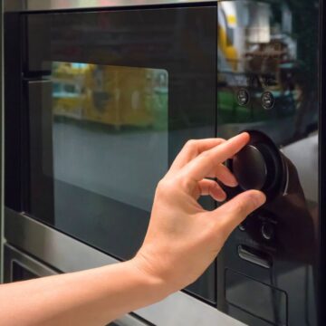 A person adjusts the settings on a microwave oven by turning a dial. The microwave is black with a reflective glass door and is positioned among other kitchen appliances. The person's hand is visible, but the rest of the body is out of frame.