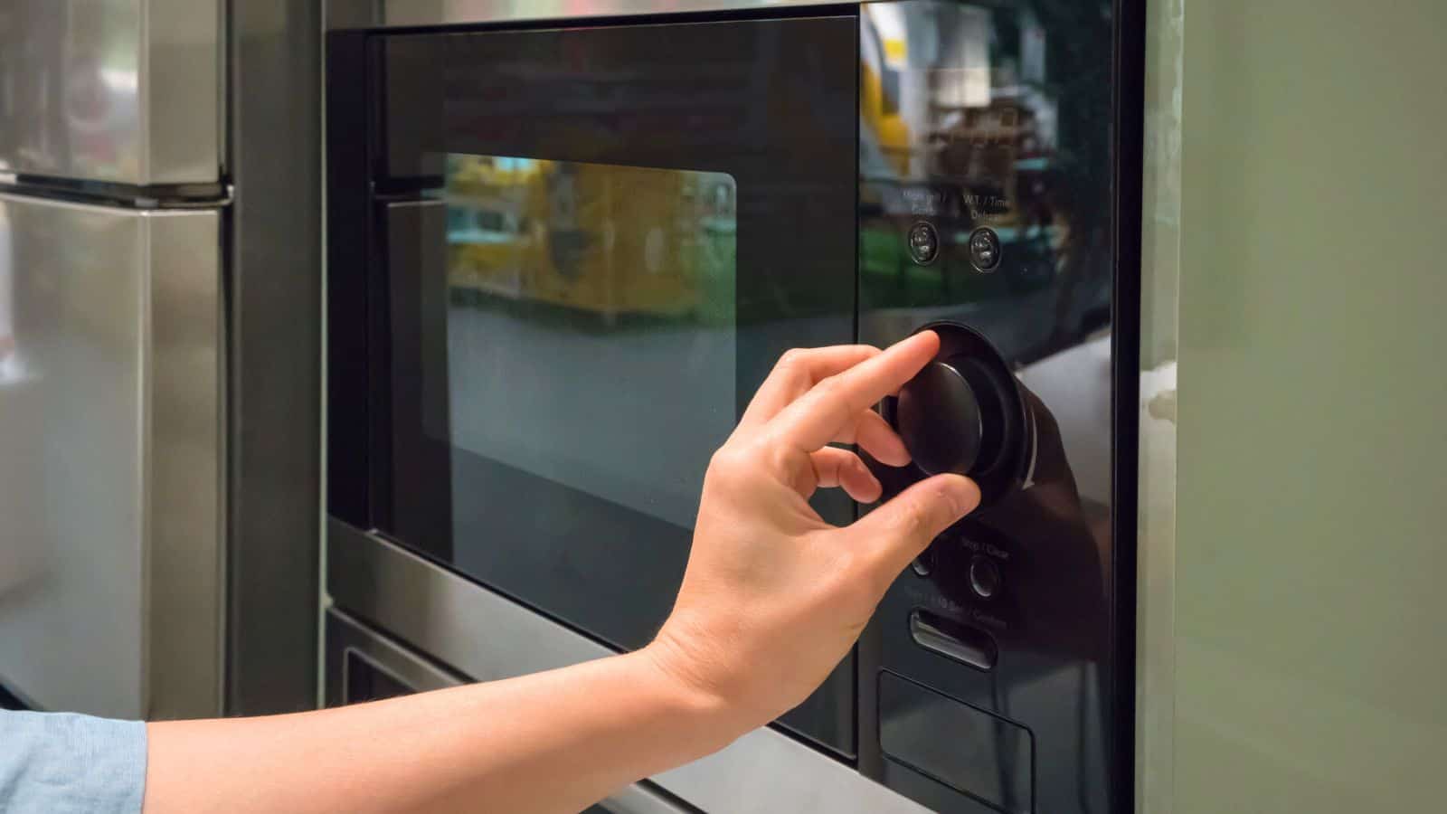 A person adjusts the settings on a microwave oven by turning a dial. The microwave is black with a reflective glass door and is positioned among other kitchen appliances. The person's hand is visible, but the rest of the body is out of frame.