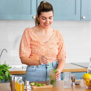 A woman in a kitchen is preparing a drink. She is stirring the drink with a straw, and fresh ingredients and utensils are on the counter. The kitchen has blue cabinets and a fruit bowl with various fruits is also visible on the counter.