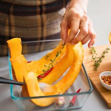 Person preparing a squash dish by placing raw squash strips into a glass container. Fresh thyme and red pepper flakes are visible, with a halved squash and a bowl of red pepper flakes on a cutting board nearby. A brush is used to coat the squash.