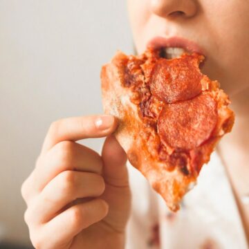 A person is taking a bite of a slice of pepperoni pizza. The pizza has a crispy, golden-brown crust and is topped with visible slices of pepperoni and melted cheese. The background is blurred, keeping the focus on the person and the pizza slice.