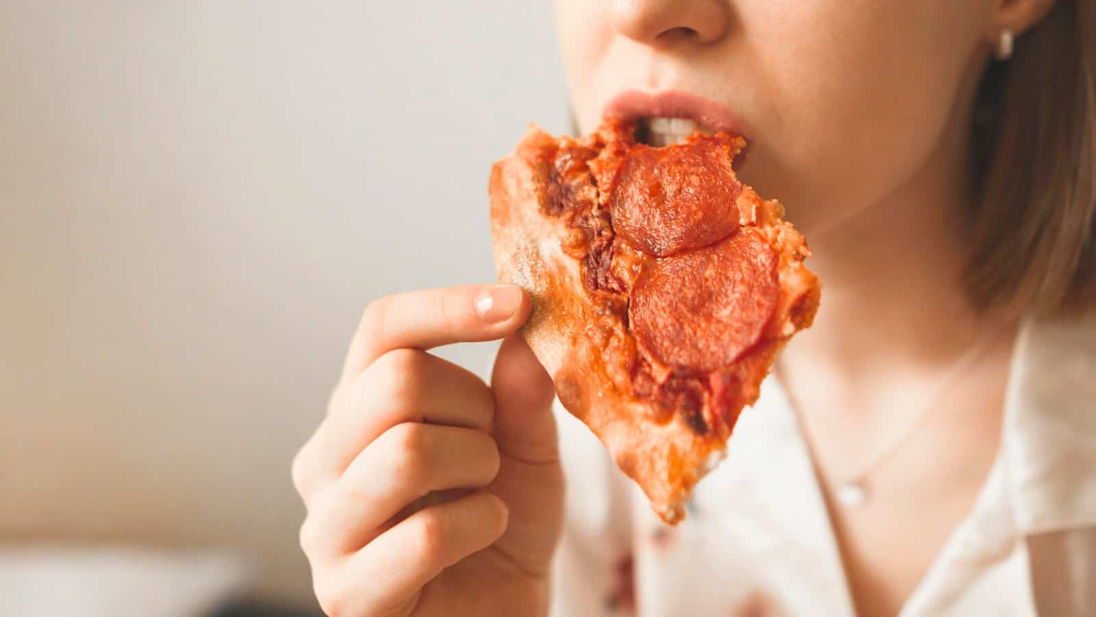 A person is taking a bite of a slice of pepperoni pizza. The pizza has a crispy, golden-brown crust and is topped with visible slices of pepperoni and melted cheese. The background is blurred, keeping the focus on the person and the pizza slice.