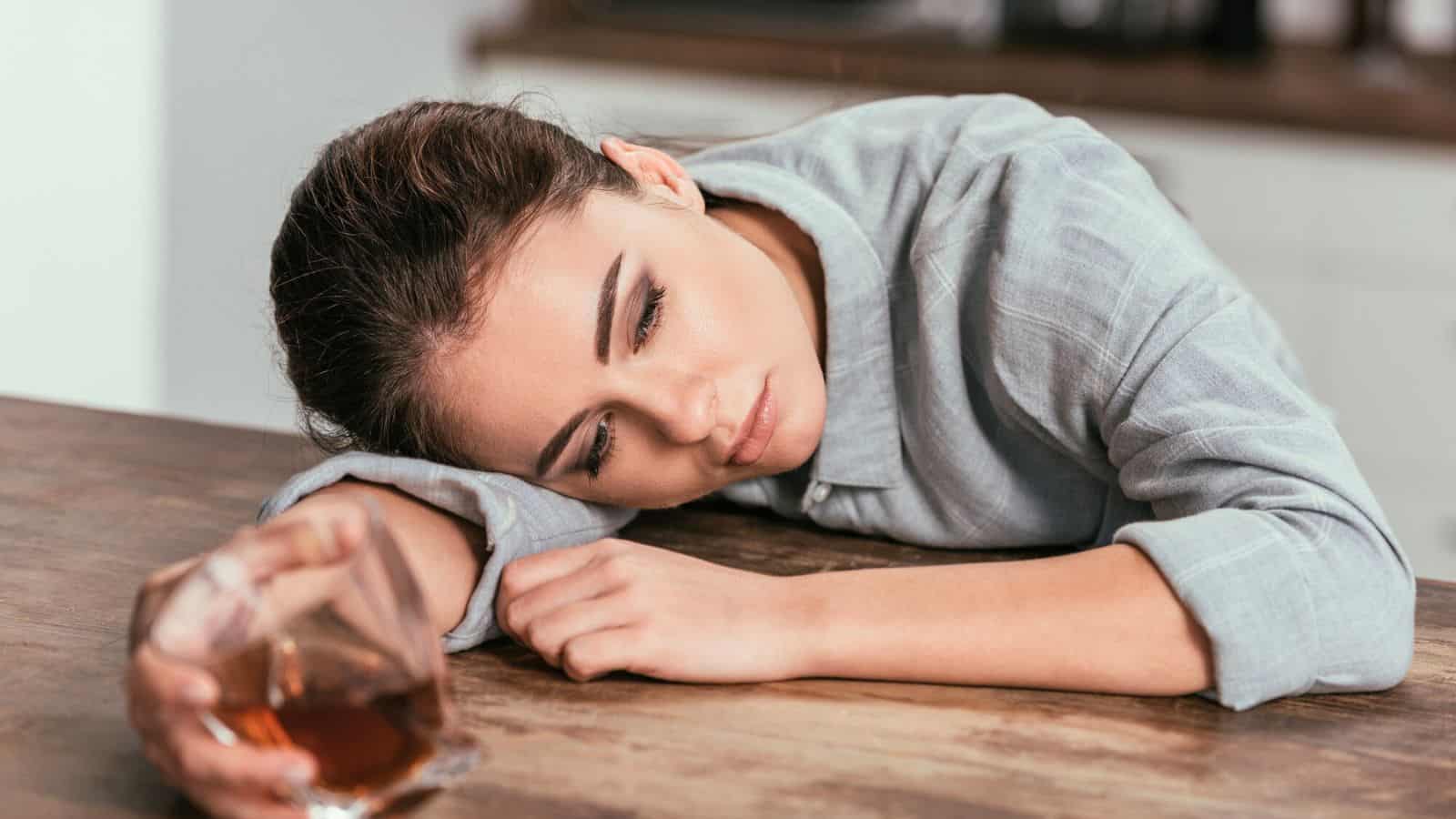 A woman is resting her head on a wooden table, her eyes closed. She is holding a glass containing an amber-colored liquid. She appears to be in a kitchen, wearing a light grey shirt.
