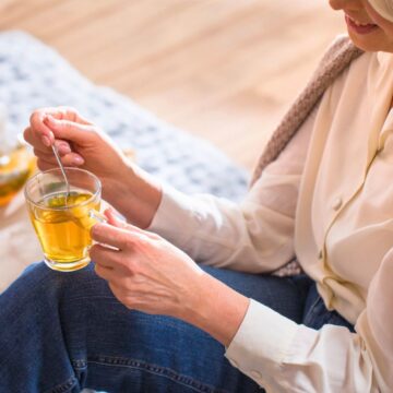 A person with grey hair is sitting, holding a cup of tea in one hand and a tea bag in the other. They are wearing a white shirt and a beige knitted cardigan. In the background, a tray with a teapot, lemon slices, and a spoon is visible on a table.