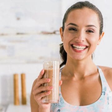 A woman smiles while holding a tall glass of a smoothie. She is wearing a floral tank top and appears to be in a kitchen setting, with a sink and various kitchen items blurred in the background.