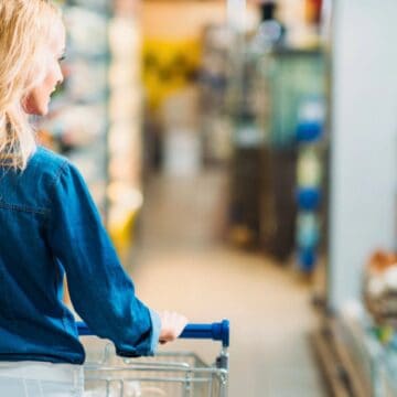A woman with blonde hair and wearing a blue denim shirt is pushing a shopping cart down an aisle in a grocery store. She is facing away from the camera, looking at the shelves stocked with various products.
