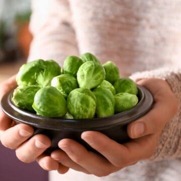 A person in a beige sweater holds a dark brown bowl filled with Brussels sprouts. The background is blurred, focusing the attention on the bowl of vegetables.