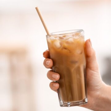 A hand holding a clear glass filled with iced coffee and ice cubes. A beige straw is placed in the drink. The background is blurred, featuring light, neutral colors.