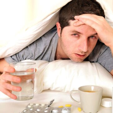 A man in bed looks unwell, with a tired expression. He is partially covered by a white blanket, holds a glass of water in one hand, and rests the other on his forehead. Nearby are a coffee mug, a bottle, and various pills on a table.
