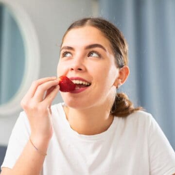 A person wearing a white shirt is eating a strawberry indoors. The background features a round mirror, light blue curtains, and a window with red frames. The person appears to be looking upward.
