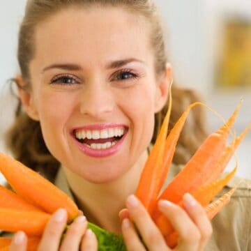 A woman holding a bunch of fresh carrots smiles at the camera. She is indoors, and the background appears to be a kitchen or similar setting.