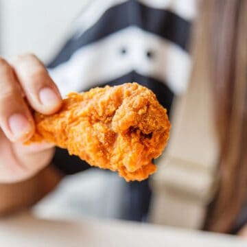 A person is holding a piece of fried chicken in their hand. The chicken has a golden-brown, crispy coating. The background is slightly blurred, but the person and their black and white shirt are partially visible.
