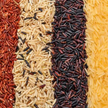 A close-up of six vertically aligned sections of different types of rice: red rice, brown rice, black rice, yellow rice, and white rice. The grains are closely packed, showing varying colors and textures.