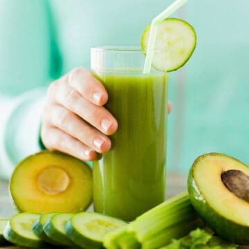 A person holds a glass of green juice with a slice of cucumber on the rim. The table in front features cut avocado halves, cucumber slices, and celery sticks. The person wears a mint sweater.