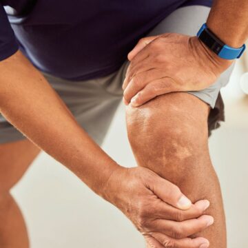 A person wearing a blue shirt and gray shorts is holding their knee with both hands, suggesting discomfort or pain. They are wearing a blue fitness tracker on their left wrist. The background is blurred.