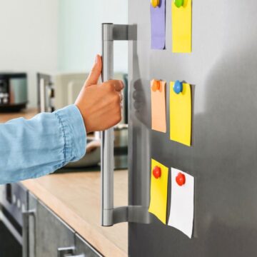 A person wearing a blue shirt opens a refrigerator door in a kitchen. The refrigerator door has colorful sticky notes attached with magnets. The kitchen counter has a coffee maker and other small appliances.
