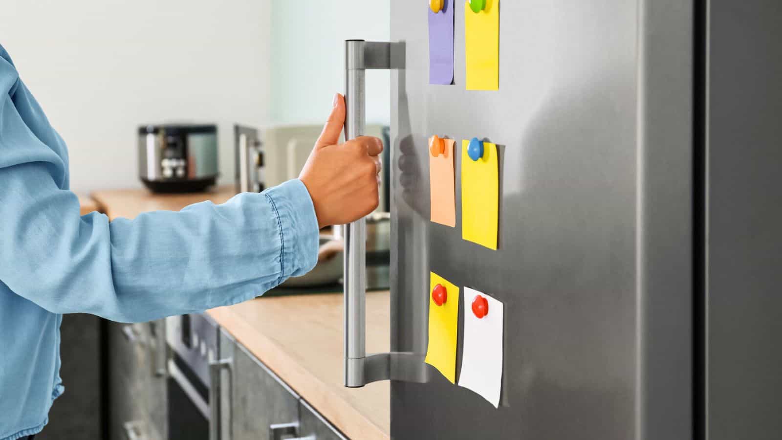 A person wearing a blue shirt opens a refrigerator door in a kitchen. The refrigerator door has colorful sticky notes attached with magnets. The kitchen counter has a coffee maker and other small appliances.
