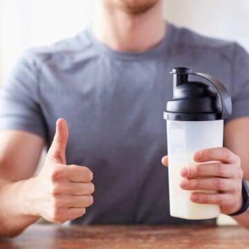 A person in a gray shirt holds a shaker bottle filled with a light-colored liquid in their right hand and gives a thumbs-up with their left hand. A large container is partially visible on the left side of the image.