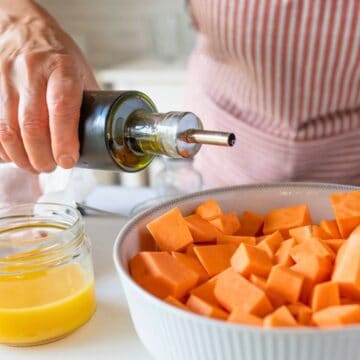 A person wearing a red and white striped apron pours olive oil from a bottle over a bowl of cubed sweet potatoes. Nearby are a bowl of raw chicken drumsticks and a small glass jar of melted butter.