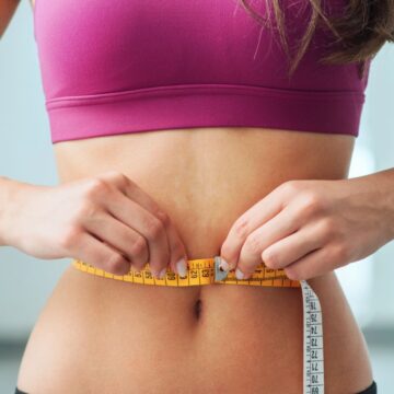 A woman in a pink sports bra measures her waist with a yellow measuring tape against a blurred background.