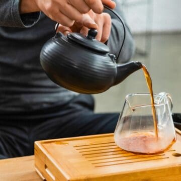 A person pours tea from a black teapot into a clear glass cup placed on a wooden tray. There are two black cups beside the tray and some steam rising from the tea. The background is blurred, focusing on the tea preparation.