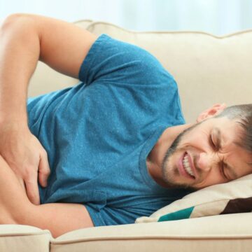 A man in a blue t-shirt lies on a beige couch grimacing in pain. He is holding his abdomen with one hand while resting his head on a cushion. The background is softly blurred, highlighting his discomfort.