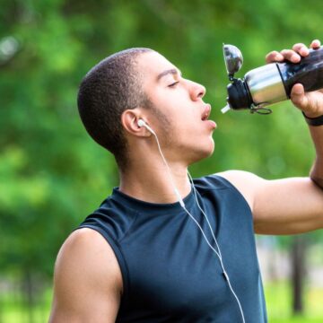 A person wearing a black sleeveless top is outdoors drinking from a water bottle. They have earphones in and a tattoo on their left forearm. Green trees are blurred in the background.