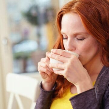 A woman with long red hair is sitting indoors and drinking from a white cup. She is wearing a gray sweater over a yellow shirt. Her eyes are closed as she enjoys her drink in a brightly lit room.
