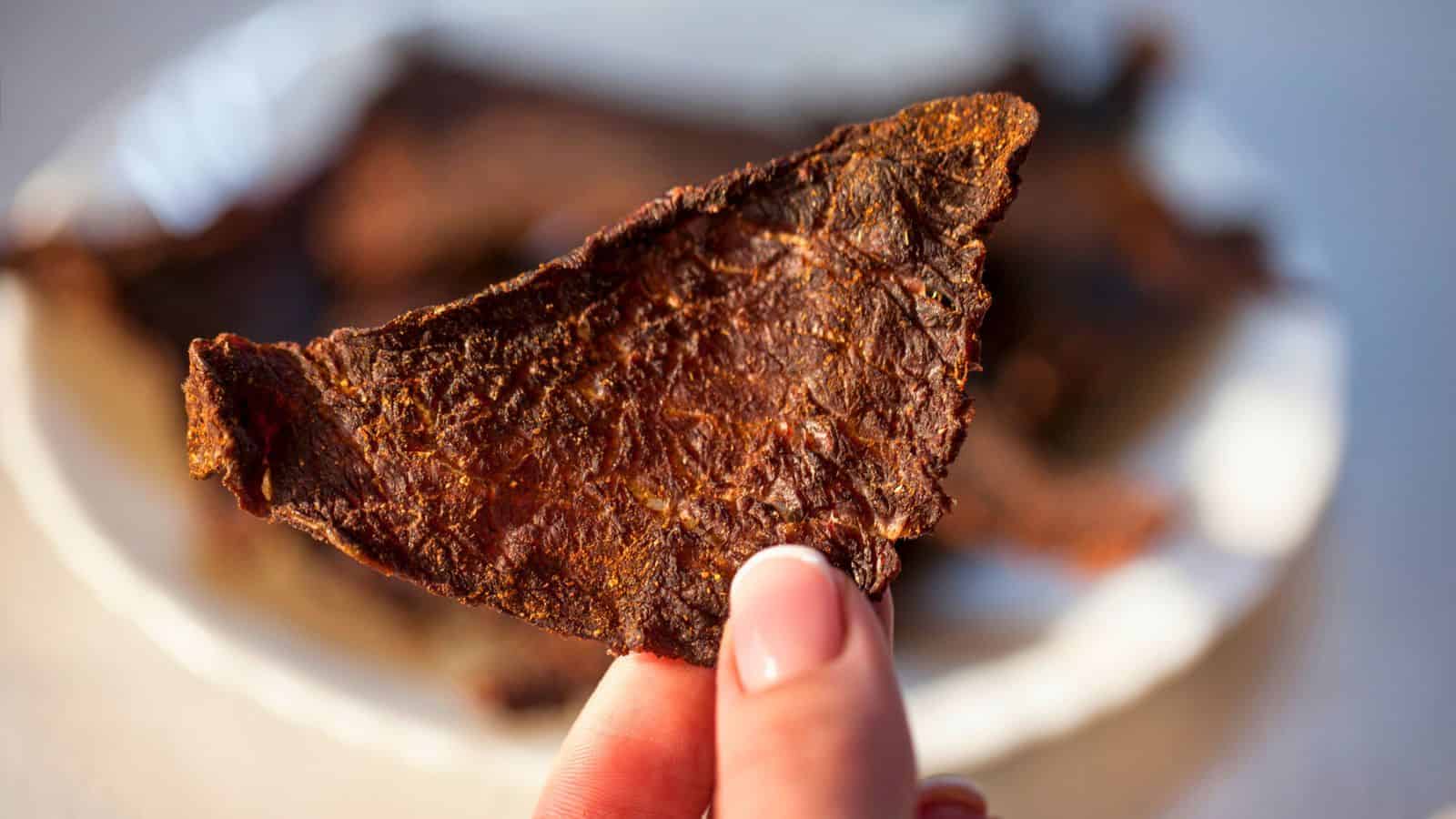 Close-up of a hand holding a piece of beef jerky. The jerky appears dark brown, textured, and slightly translucent, with a plate of more jerky pieces blurred in the background. The image highlights the detail and texture of the beef jerky.