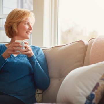 A woman sits on a sofa holding a mug, looking out the window with a relaxed expression. She is wearing a blue long-sleeve shirt and has short blonde hair. There are several cushions on the sofa, and sunlight is streaming through the window.