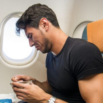 A man with dark hair and a beard sits in an airplane seat by a window, holding a small container of food in his hands. He is wearing a black t-shirt and appears focused on the food. The seat has a striped blue and orange upholstery.