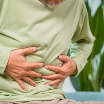 A person wearing a green long-sleeve shirt is sitting and clutching their stomach with both hands, indicating discomfort or pain. Large green leaves are visible in the background. The person's face is not shown in the image.