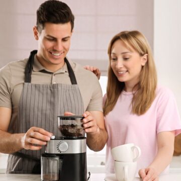 A man and a woman are in a kitchen using a coffee grinder. The man, wearing an apron, is operating the grinder, while the woman holds a white coffee mug and smiles. Various kitchen items are visible in the background, including jars and a towel.