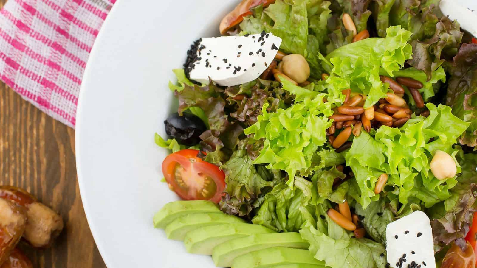 A bowl of salad with a variety of ingredients including leafy greens, cherry tomatoes, sliced avocado, cubes of white cheese topped with black sesame seeds, nuts, and seeds. The bowl is placed on a wooden table with a red-checked cloth partially visible.