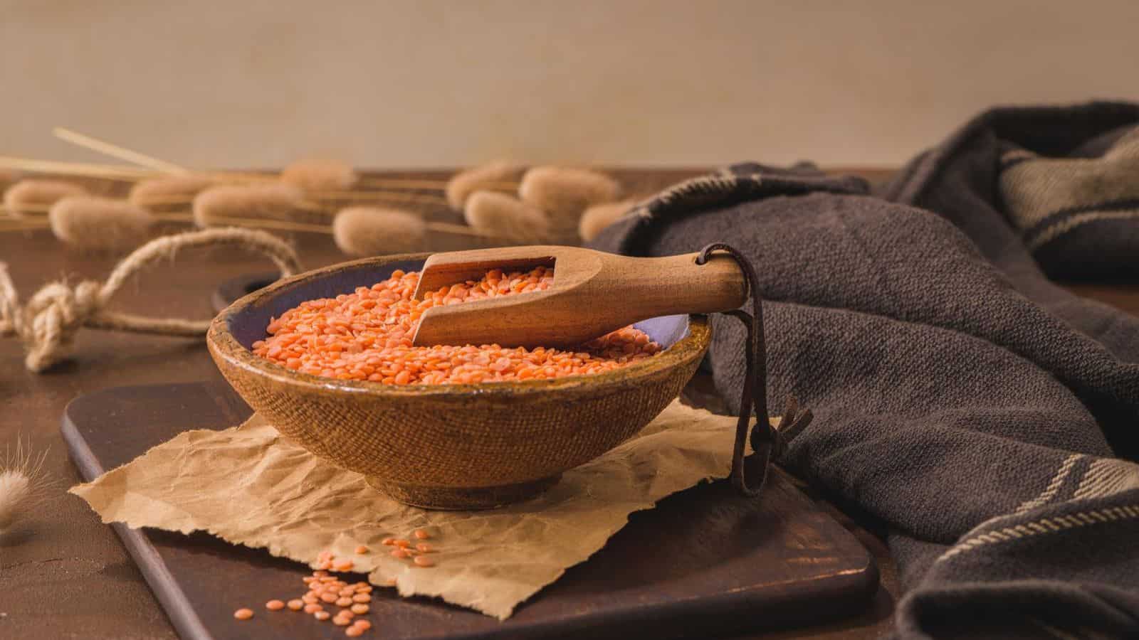 A wooden bowl filled with red lentils sits on a piece of parchment paper. A wooden scoop rests in the lentils. The bowl is placed on a dark wooden surface with dried pampas grass and a folded cloth in the background.