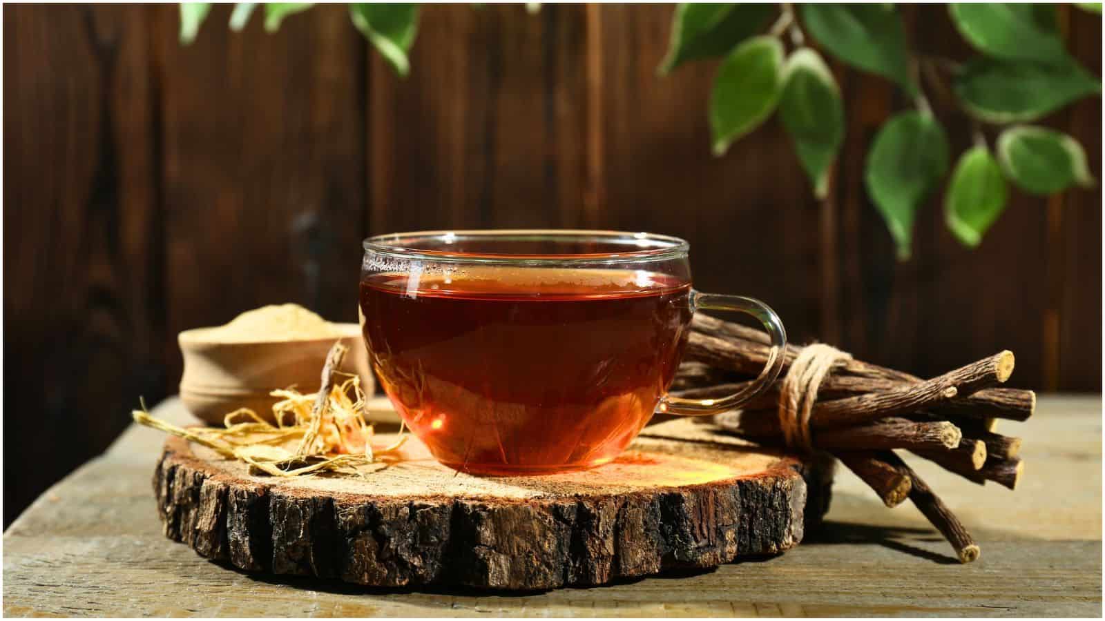 A clear glass cup filled with tea sits on a rustic wooden coaster. Nearby, there are bundles of dried herbs tied together with string, and a small bowl filled with a powdered substance. Green leaves hang in the top right corner of the image.