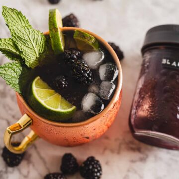 An iced beverage in a copper mug garnished with mint leaves, a lime slice, and blackberries sits on a marble surface. Nearby is a jar labeled with "BLACKB" containing a blackberry mixture. Loose blackberries are scattered around, creating an enticing scene that looks fresh off an auto draft.