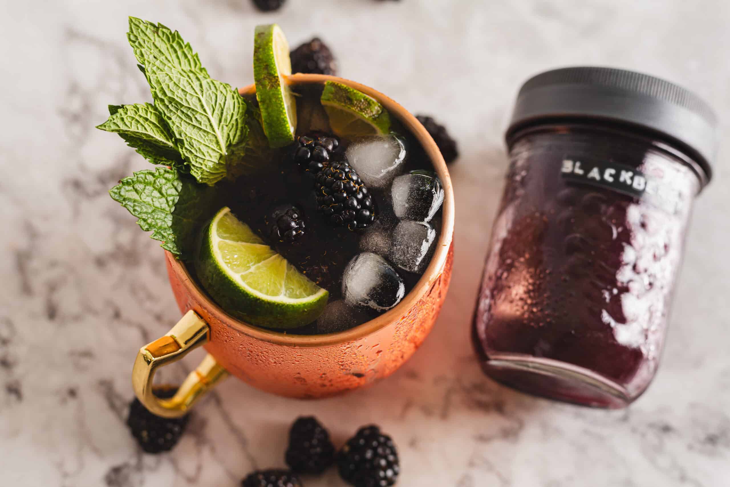 An iced beverage in a copper mug garnished with mint leaves, a lime slice, and blackberries sits on a marble surface. Nearby is a jar labeled with "BLACKB" containing a blackberry mixture. Loose blackberries are scattered around, creating an enticing scene that looks fresh off an auto draft.