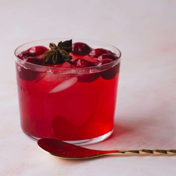 A glass filled with a red drink containing ice cubes and what appear to be cherries is placed on a light-colored surface. The drink, perfect for fall recipes, is garnished with star anise. A twisted gold spoon rests beside the glass, ready to sweeten up your day.