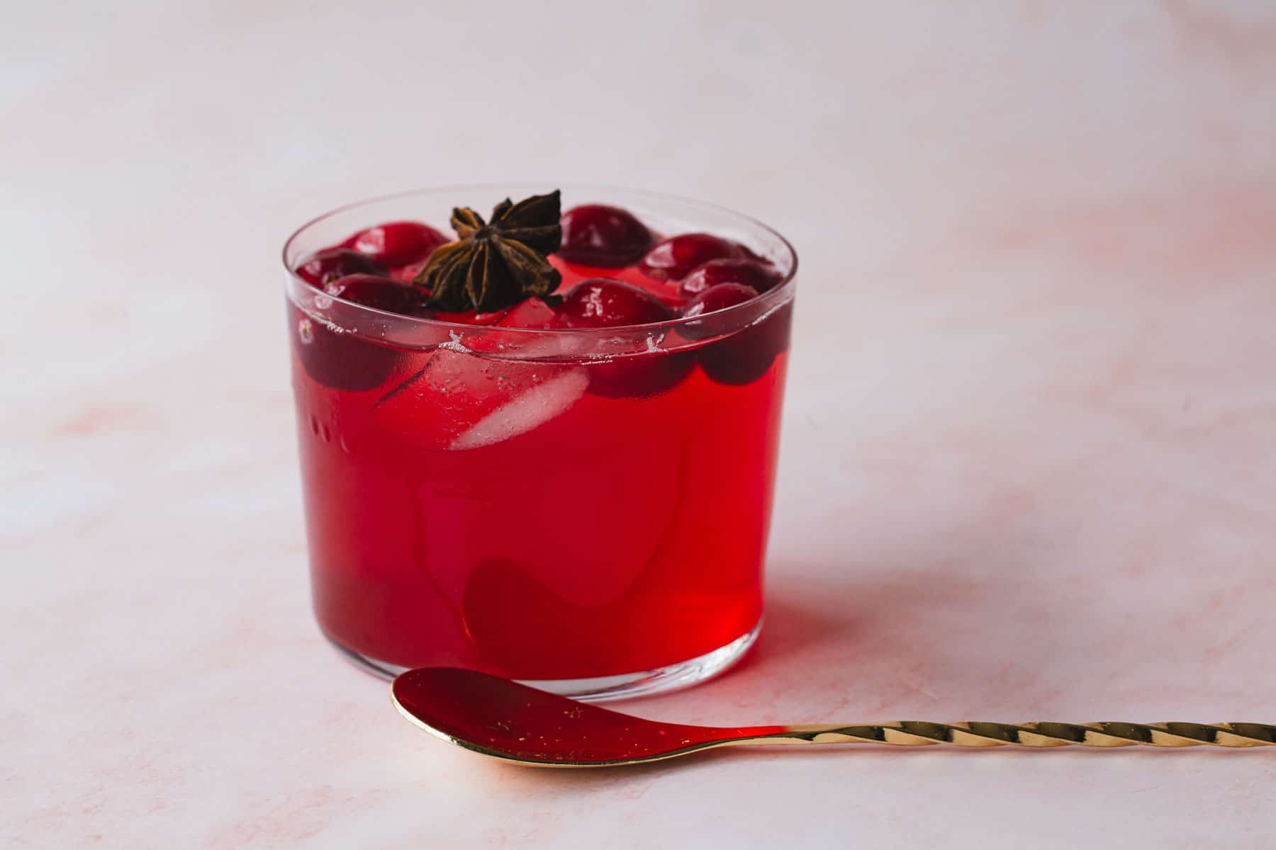 A glass filled with a red drink containing ice cubes and what appear to be cherries is placed on a light-colored surface. The drink, perfect for fall recipes, is garnished with star anise. A twisted gold spoon rests beside the glass, ready to sweeten up your day.