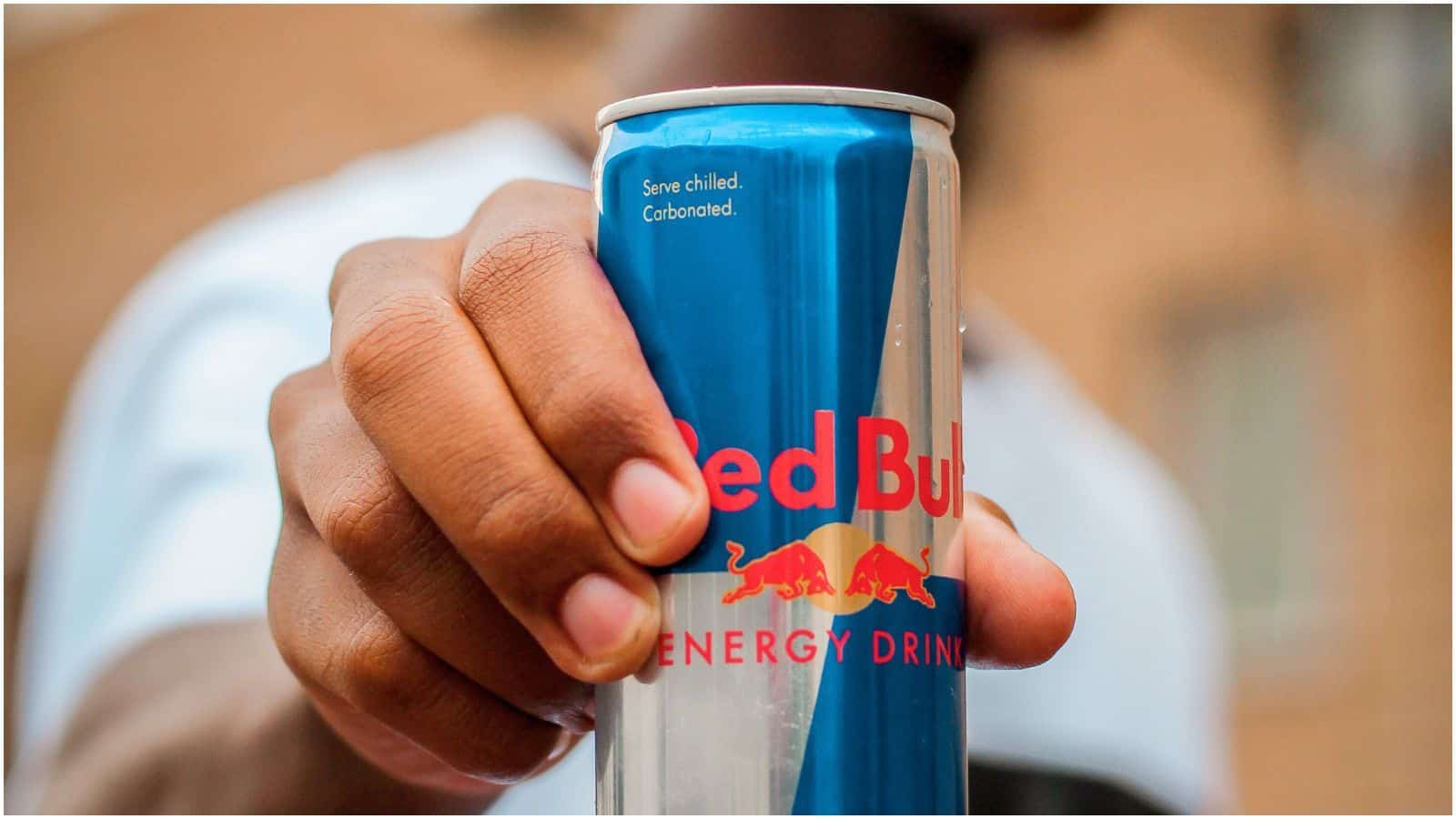 A person is holding a can of Red Bull energy drink. The can is primarily blue and silver, with a red and yellow logo on the front. The person's hand is prominently featured in the foreground, while their face is out of focus in the background.