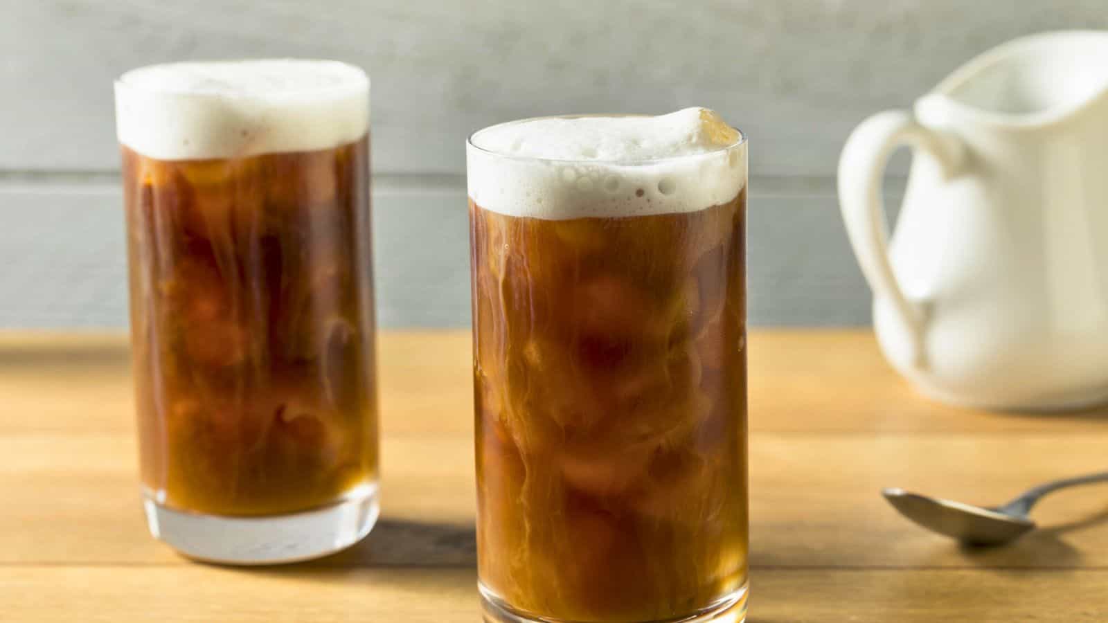 Two tall glasses of iced cold brew coffee with frothy tops are placed on a wooden surface. An empty white pitcher and a spoon lie in the background on the right side. The background is a light-colored, textured wall.