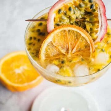 A cocktail garnished with a slice of orange and half of a passion fruit is placed on a marble surface. Behind the glass, there is an additional half of an orange. The drink contains visible ice cubes and passion fruit seeds, creating a delightful fusion that’s anything but an Auto Draft.
