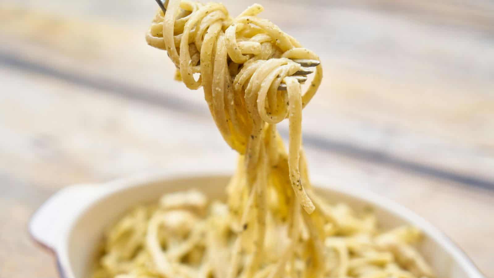 A close-up image of a fork holding a twirl of creamy pasta over a bowl filled with more pasta. The background is blurred, highlighting the pasta’s texture and sauce.