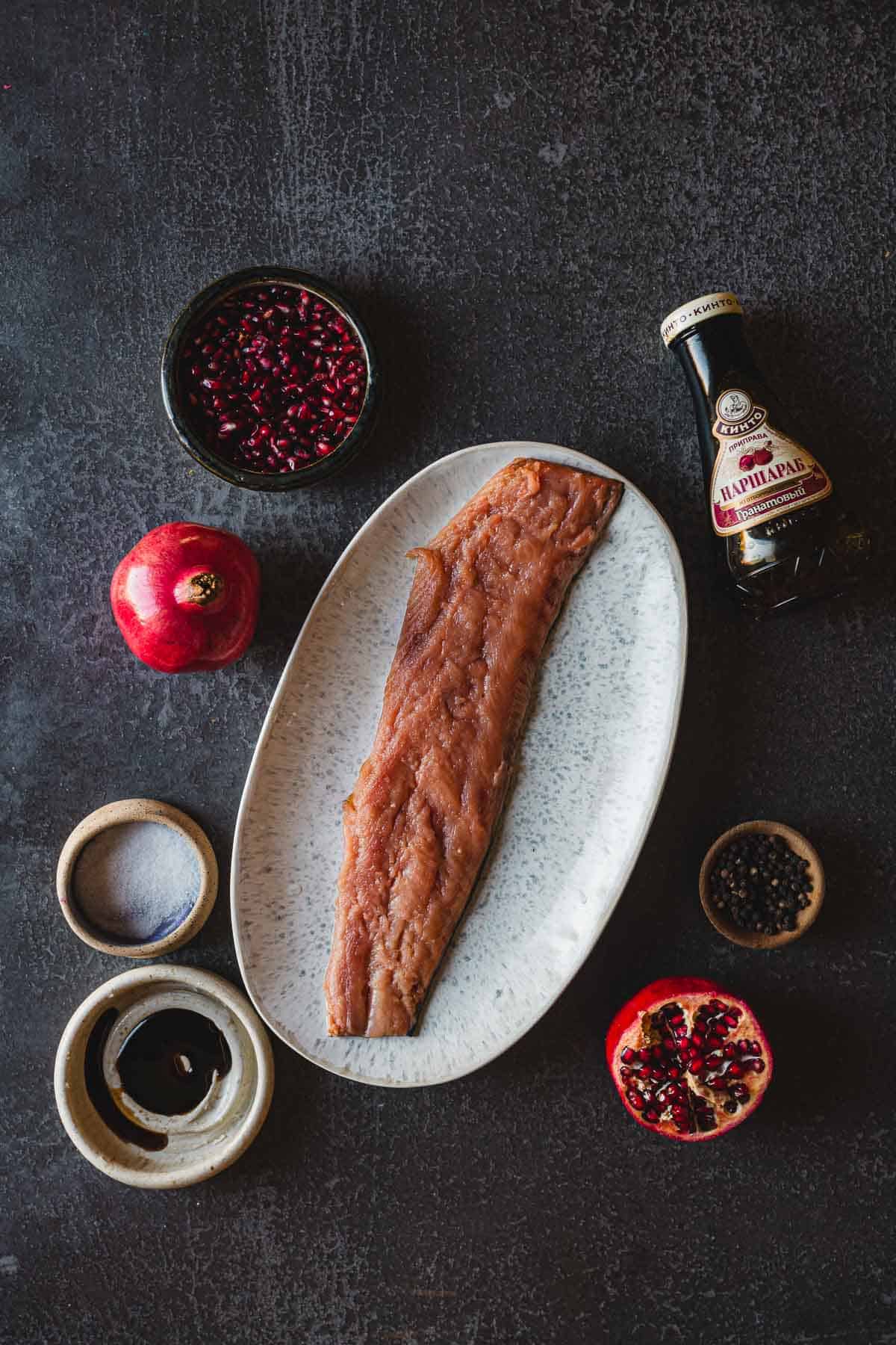 Top view of a piece of raw fish fillet on a white oval plate surrounded by ingredients, including pomegranate, halved pomegranate, a small bowl of pomegranate seeds, a bottle of pomegranate molasses, a bowl of salt, sauce, and peppercorns—a perfect setup for Pomegranate Glazed Salmon.