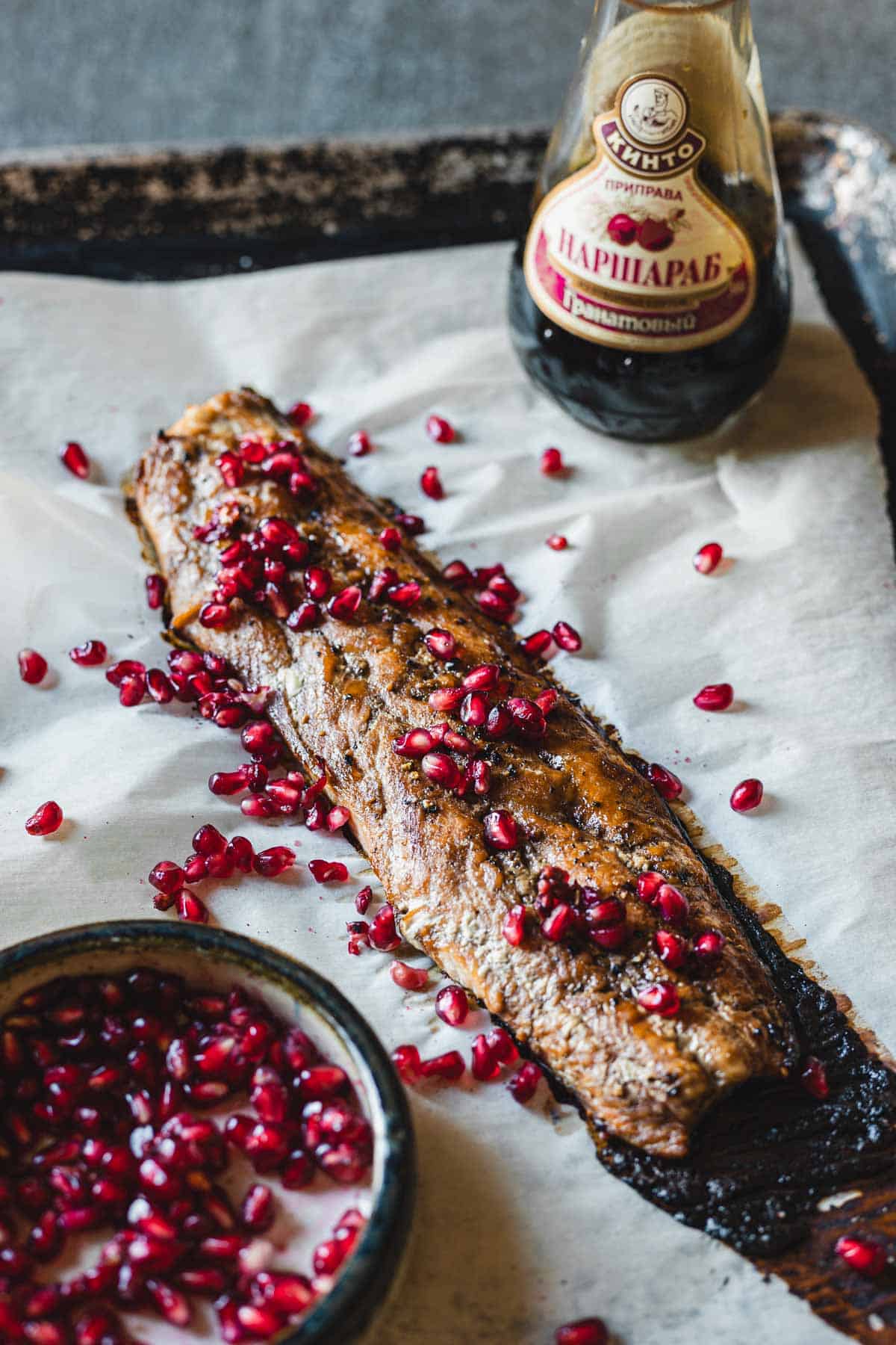 A grilled eggplant topped with pomegranate seeds placed on parchment paper. A bowl of pomegranate seeds sits to the side, and a bottle of Pomegranate Glazed Sauce "Narsharab" is partially visible in the background, enhancing this elegant dish reminiscent of an oven-baked masterpiece.