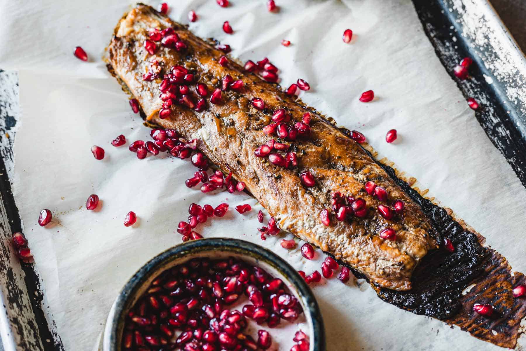 A pomegranate glazed salmon fillet is placed on parchment paper in a baking tray, garnished with pomegranate seeds scattered over and around it. A small bowl with additional pomegranate seeds is positioned in the lower left corner of the tray.