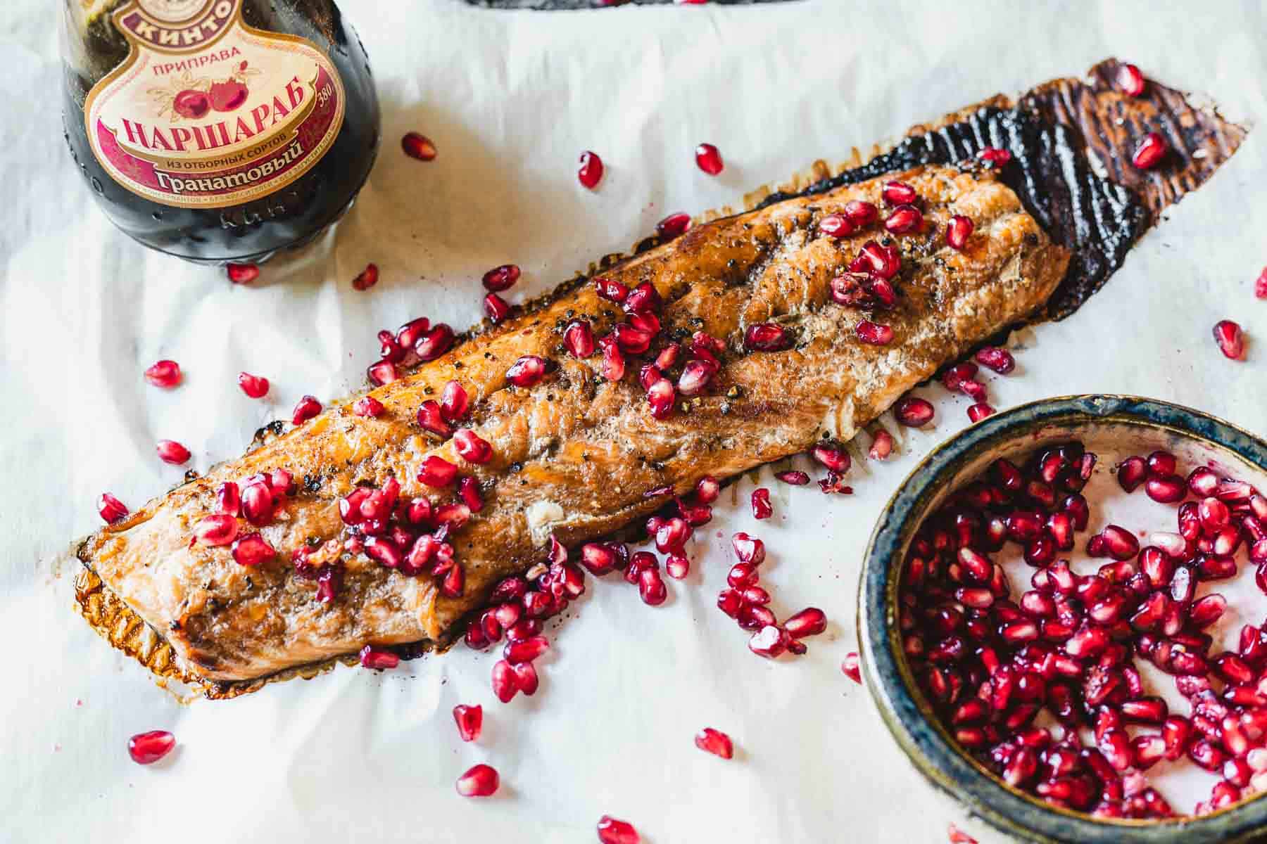 Pomegranate glazed salmon fillet garnished with pomegranate seeds on parchment paper, accompanied by a jar of marinade and a bowl of additional pomegranate seeds.