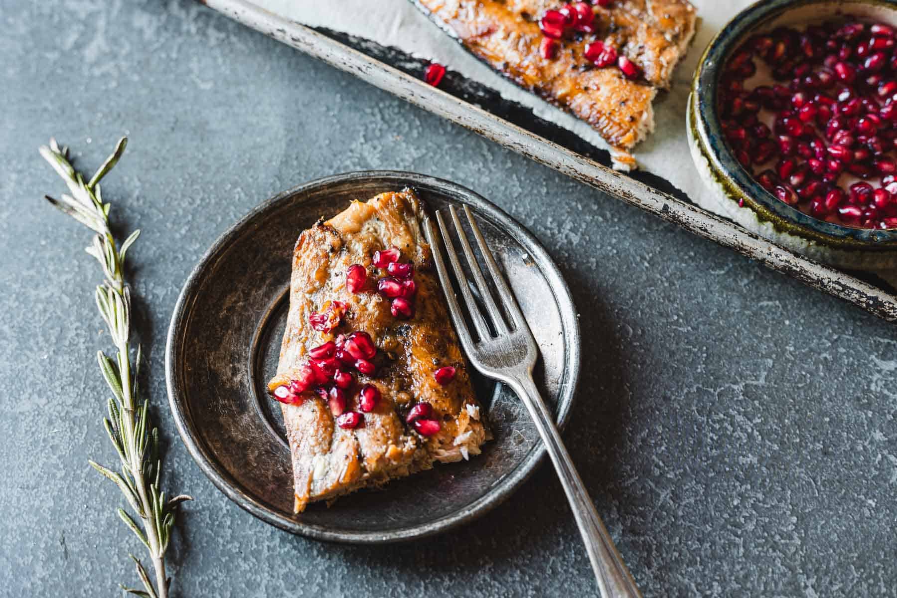 A plate with a piece of pomegranate glazed salmon garnished with pomegranate seeds and a fork. In the background, another piece of fish on a tray and a small bowl with pomegranate seeds can be seen. A sprig of rosemary is placed next to the plate.